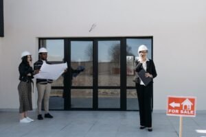 Professionnels en casques d'architecte devant un bâtiment avec panneau "À vendre". Étudient des plans
