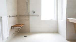Salle de bain accessible senior, douche de plain-pied avec barre d'appui et siège, carrelage clair, lumineuse
