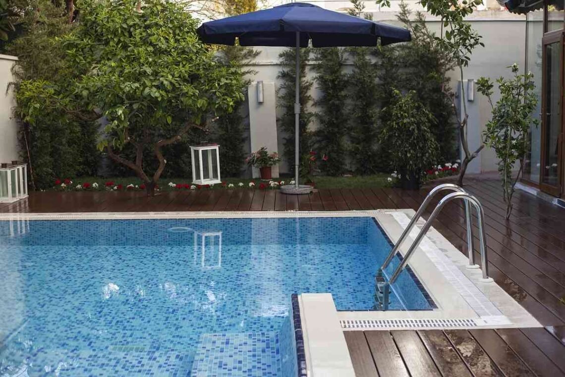 Une piscine extérieure avec terrasse en bois, parasol bleu et végétation