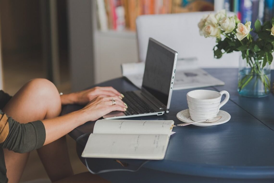 Une personne travaille sur ordinateur, note dans cahier, boit café, fleurs à côté