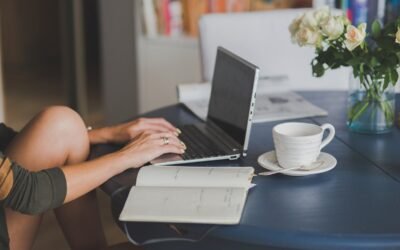 Une personne travaille sur ordinateur, note dans cahier, boit café, fleurs à côté