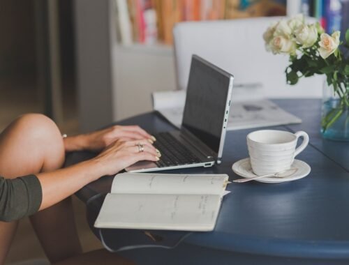 Une personne travaille sur ordinateur, note dans cahier, boit café, fleurs à côté