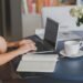 Une personne travaille sur ordinateur, note dans cahier, boit café, fleurs à côté